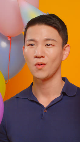 Vertical-Video-Studio-Portrait-Of-Man-Wearing-Party-Hat-Celebrating-Birthday-Hiding-Behind-Balloons